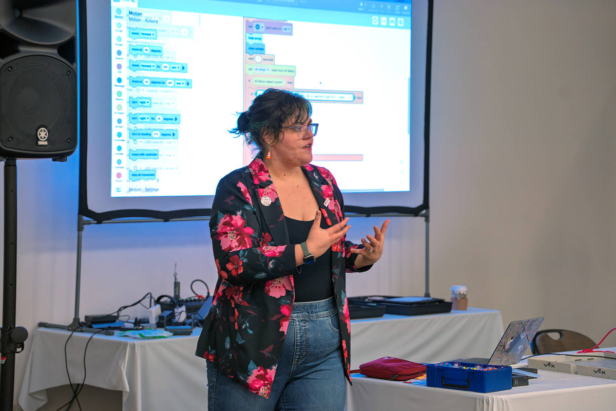 A woman wearing glasses, a black top, and a black floral blazer with pink flowers, standing in front of a projector screen showing a coding interface. She is in a presentation room with a speaker on the left and tables with equipment below.