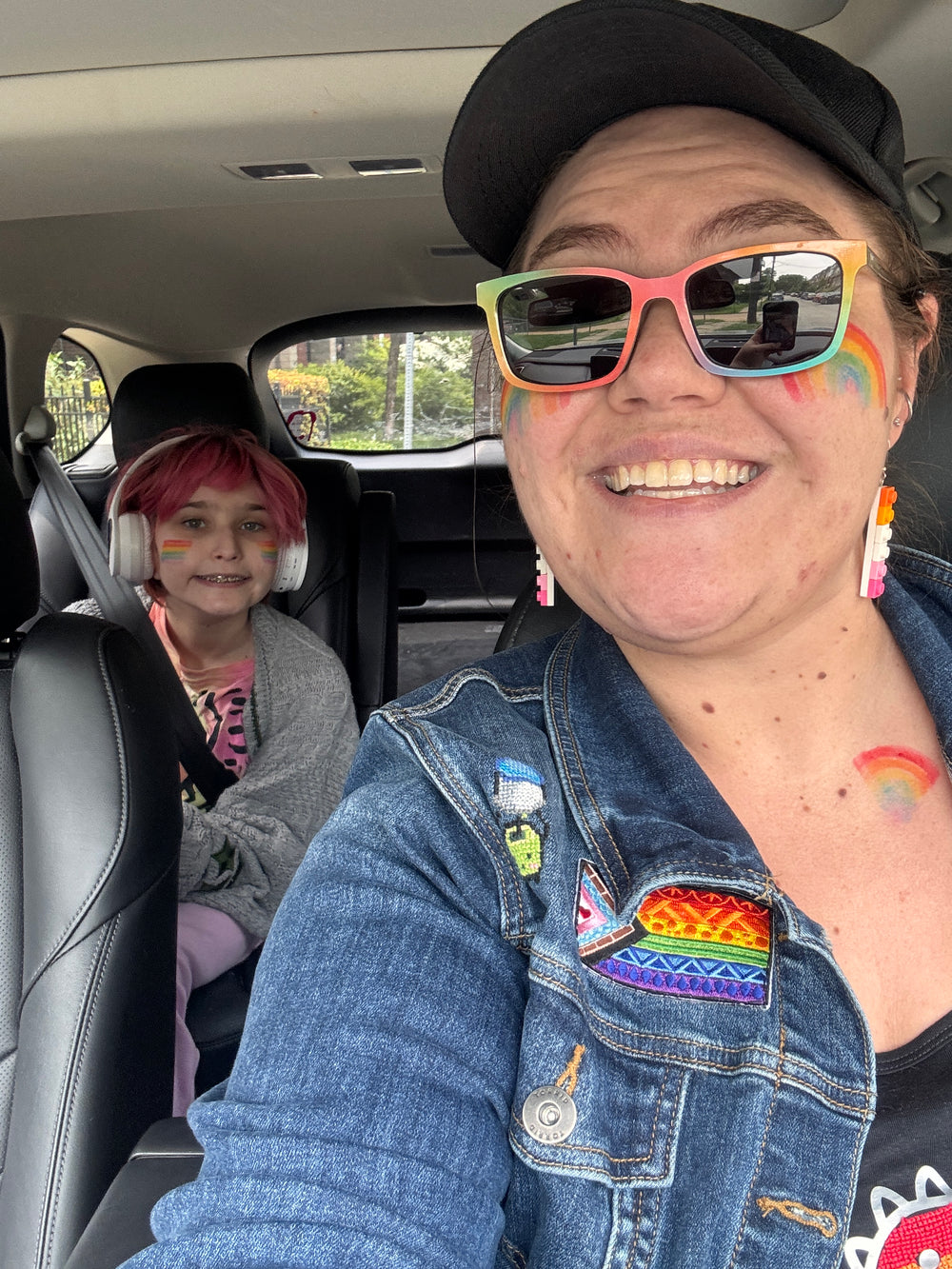 A smiling woman in a black cap and colorful rainbow sunglasses takes a selfie inside a car with a child in the back seat wearing pink hair and white headphones. The woman wears a denim jacket with rainbow-themed patches and a rainbow tattoo on her neck.