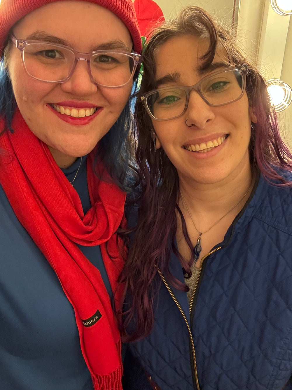 Two women smiling for a selfie in a brightly lit dressing room. The woman on the left wears a red beanie, pink-framed glasses, a red scarf, and a blue top.