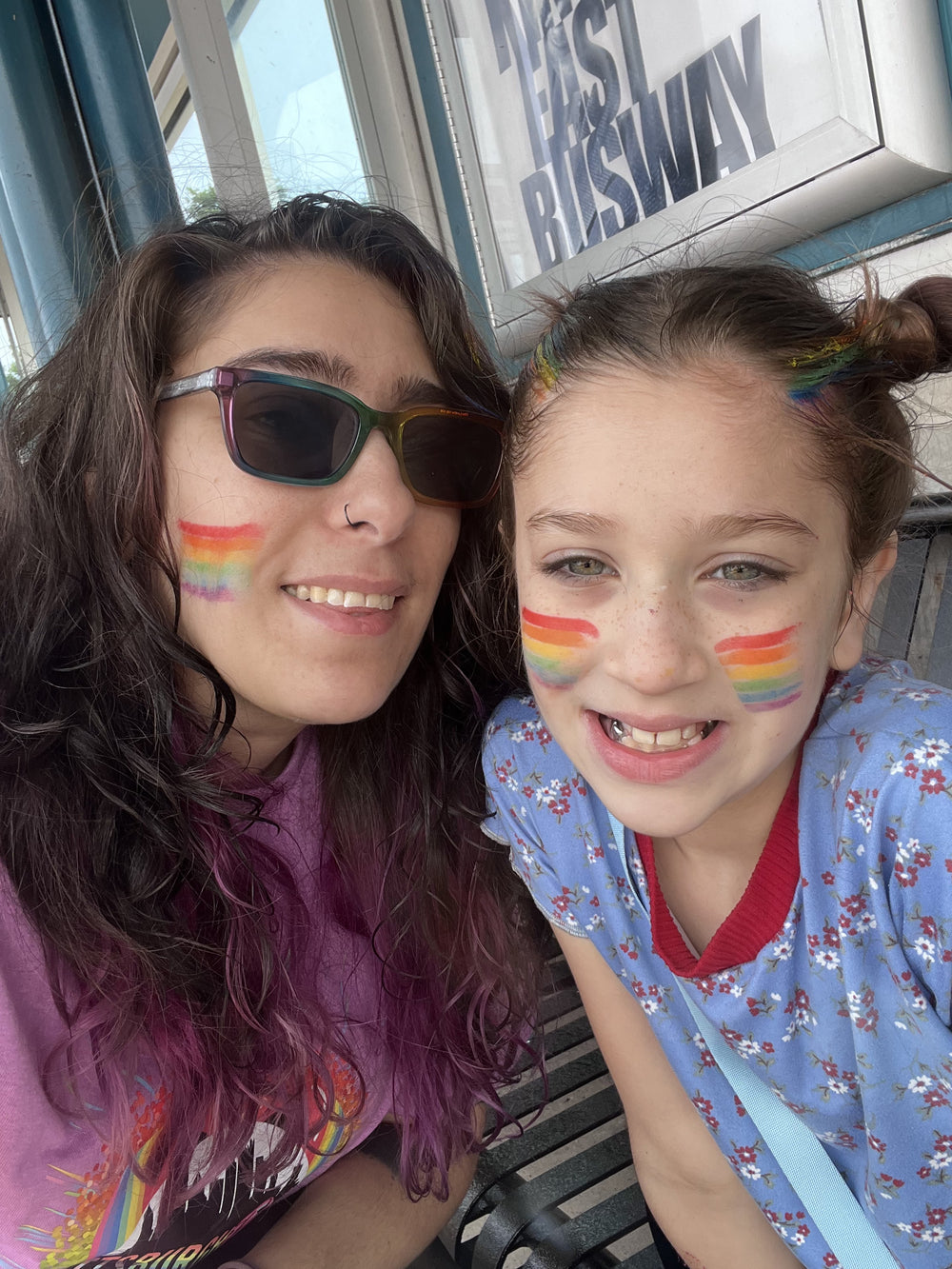 Two young women smiling and taking a selfie on a bus, both wearing rainbow face paint on their cheeks. The woman on the left wears a purple graphic t-shirt and sunglasses, while the girl on the right wears a blue floral shirt.