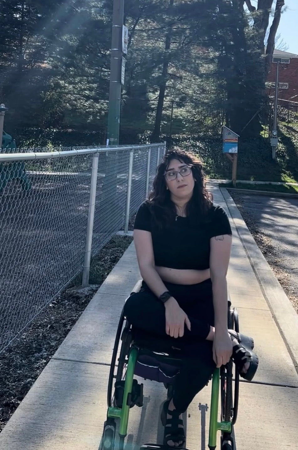 A person with dark curly hair and glasses sitting in a green manual wheelchair on an outdoor concrete path, wearing a black crop top and black pants.