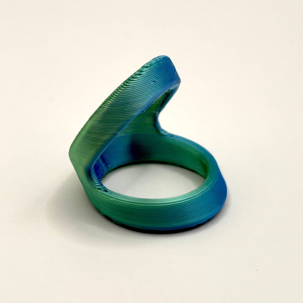 A 3D-printed ring with a unique spiral design, featuring a blue and green gradient finish, photographed on a neutral gray background.