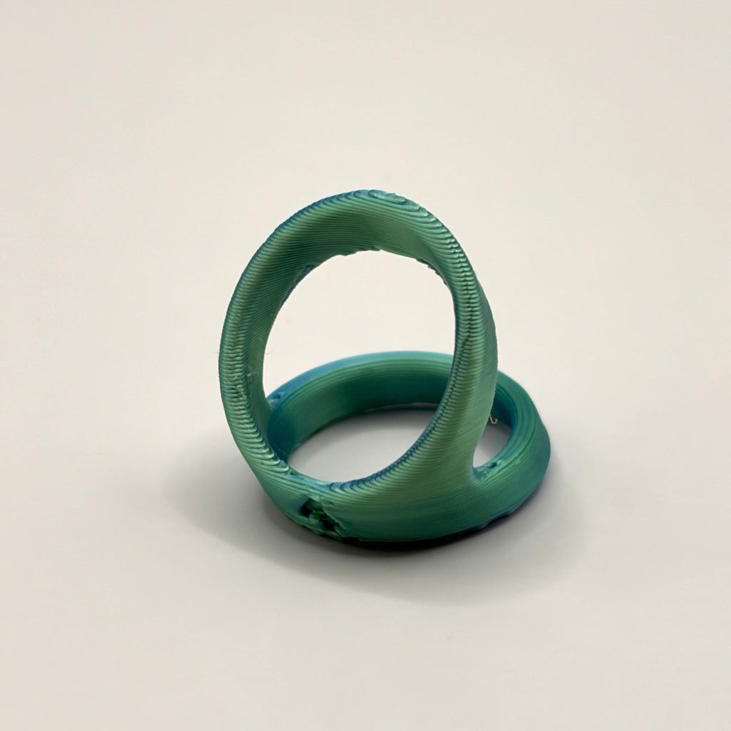 A 3d printed ring n a metallic teal green color, displayed on a neutral light gray background.