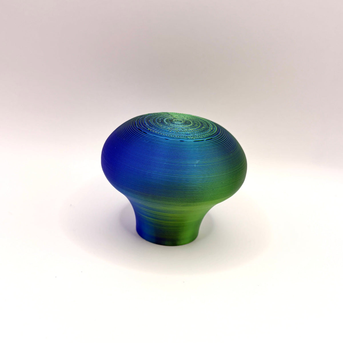 A small decorative object with a rounded, mushroom-like shape featuring a blue-to-green gradient and subtle spiral detailing on top, set against a plain white background.