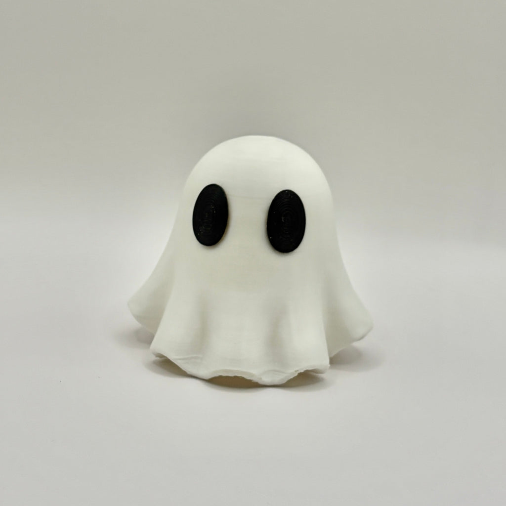 A small white ghost figurine with large black oval eyes, standing on a light gray surface against a light gray background.