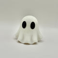 A small white ghost figurine with large black oval eyes, standing on a light gray surface against a light gray background.