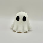 A small white ghost figurine with large black oval eyes, standing on a light gray surface against a light gray background.