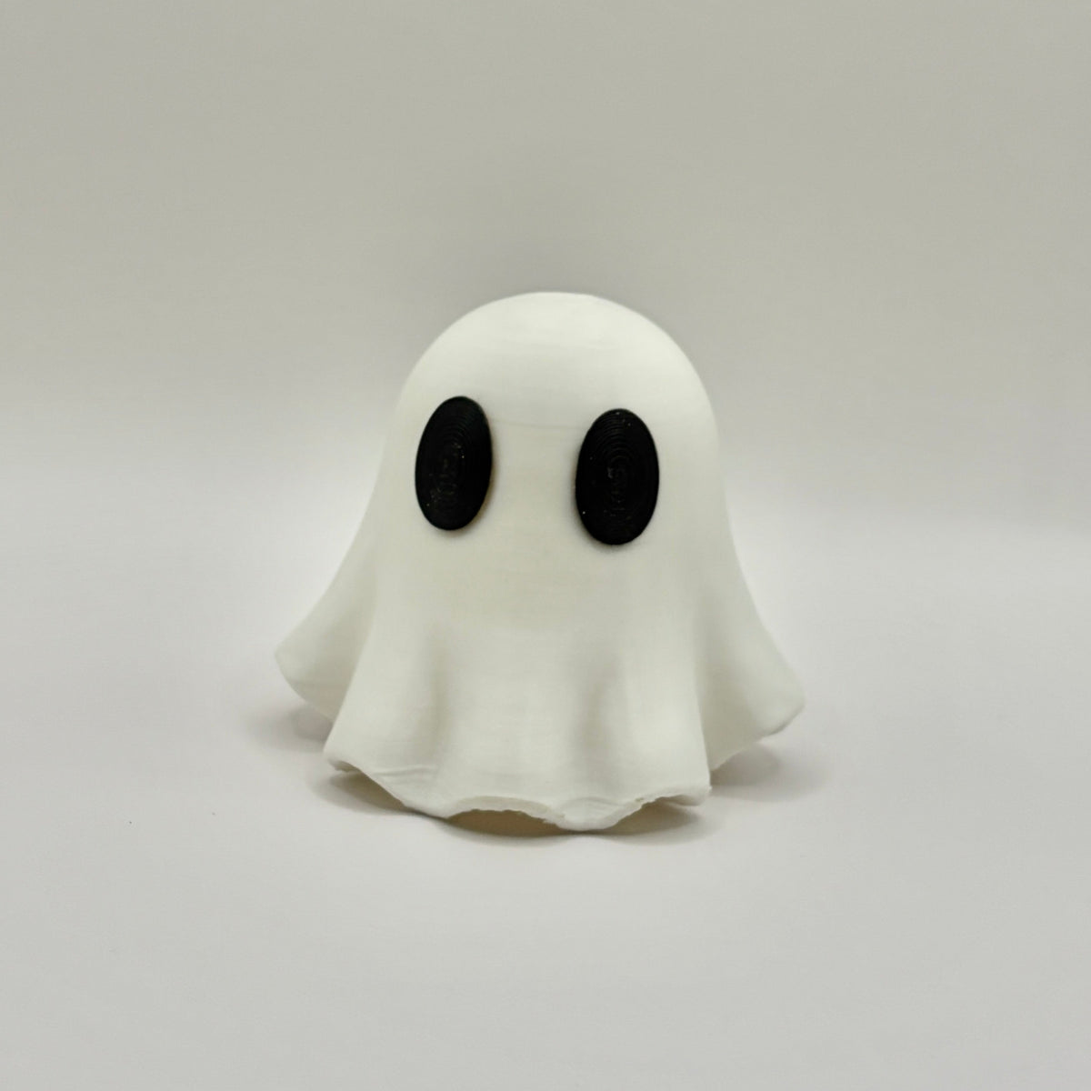 A small white ghost figurine with large black oval eyes, standing on a light gray surface against a light gray background.