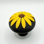 A round black knob featuring a raised sunflower flower design on top, set against a plain white background.