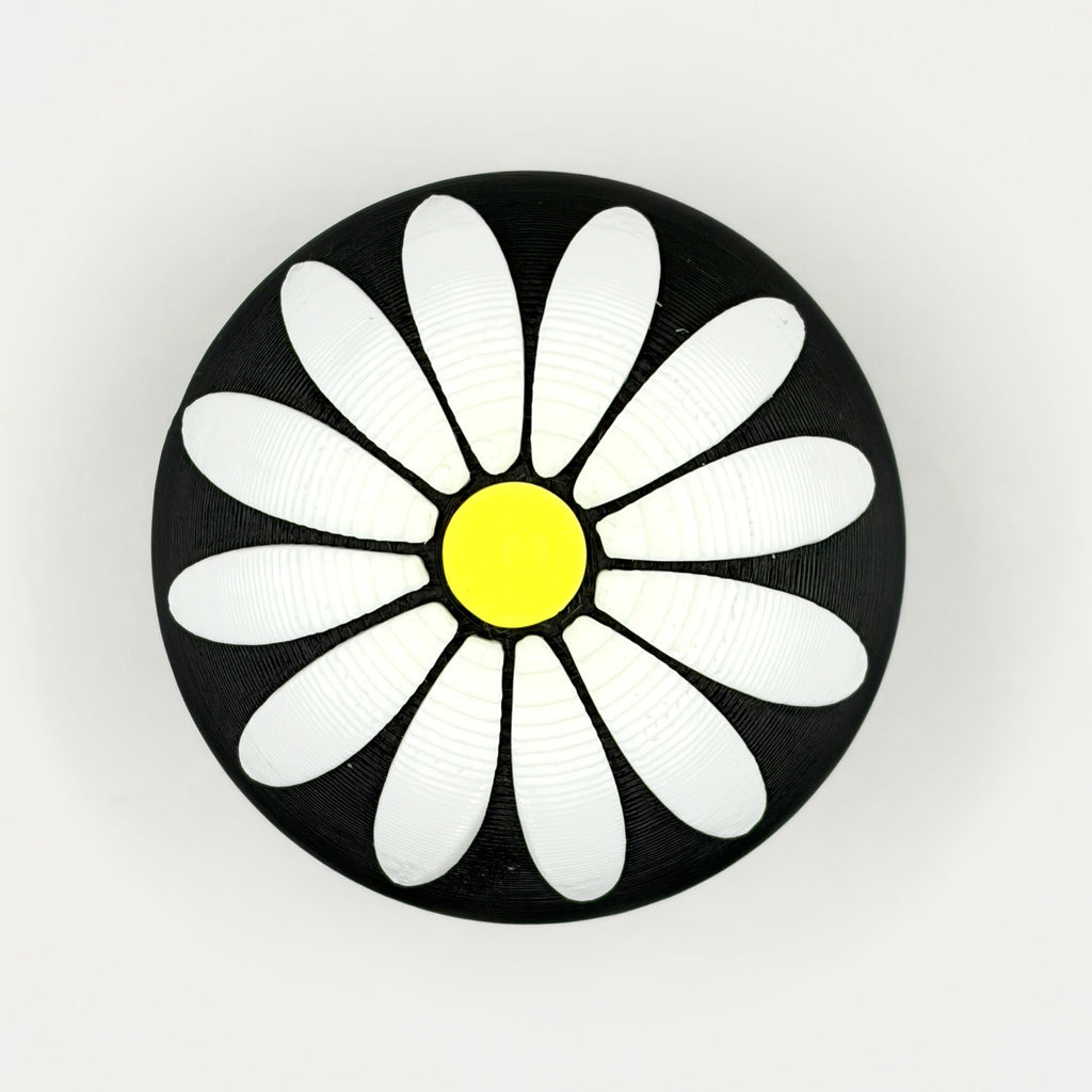 A round black button or badge featuring a stylized white daisy flower with a bright yellow center, set against a plain light gray background.