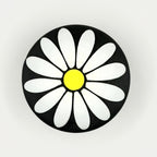 A round black button or badge featuring a stylized white daisy flower with a bright yellow center, set against a plain light gray background.