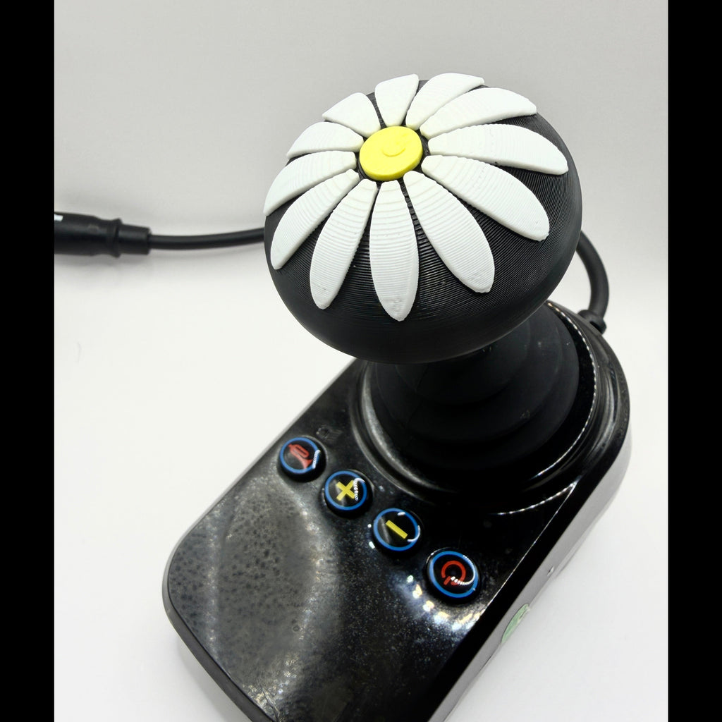 A black joystick controller with a daisy pattern on the knob, featuring white petals, a yellow center, and four blue-bordered buttons on the base, shown on a light gray background.