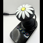 A black joystick controller with a daisy pattern on the knob, featuring white petals, a yellow center, and four blue-bordered buttons on the base, shown on a light gray background.
