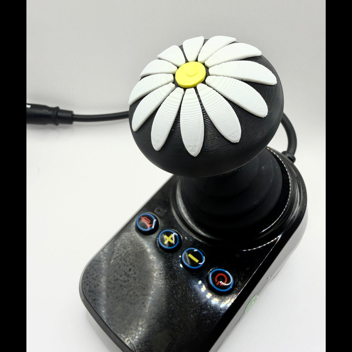 A black joystick controller with a daisy pattern on the knob, featuring white petals, a yellow center, and four blue-bordered buttons on the base, shown on a light gray background.