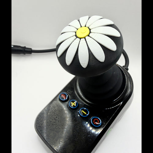 A black joystick controller with a daisy pattern on the knob, featuring white petals, a yellow center, and four blue-bordered buttons on the base, shown on a light gray background.
