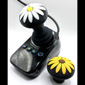 A black joystick controller with a daisy flower knob on top, shown alongside a matching black knob featuring yellow flower petals and a brown center, on a white background.