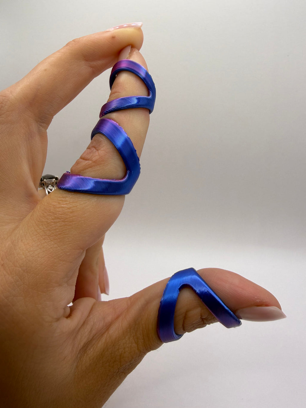 A close-up of a hand wearing three iridescent blue and purple satin finger rings, with a silver band on the middle finger, against a plain light gray background.