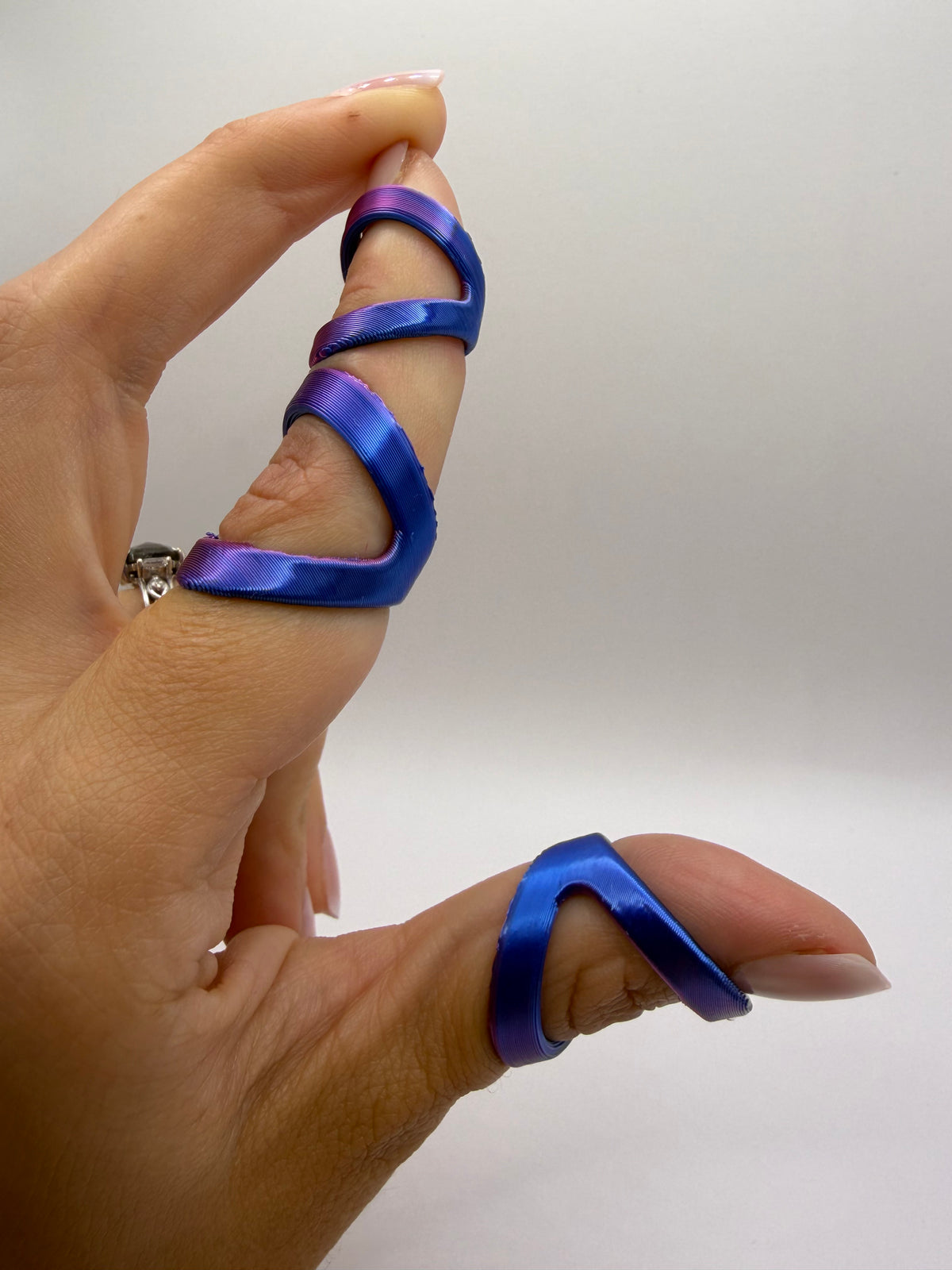 A close-up of a hand wearing three iridescent blue and purple satin finger rings, with a silver band on the middle finger, against a plain light gray background.