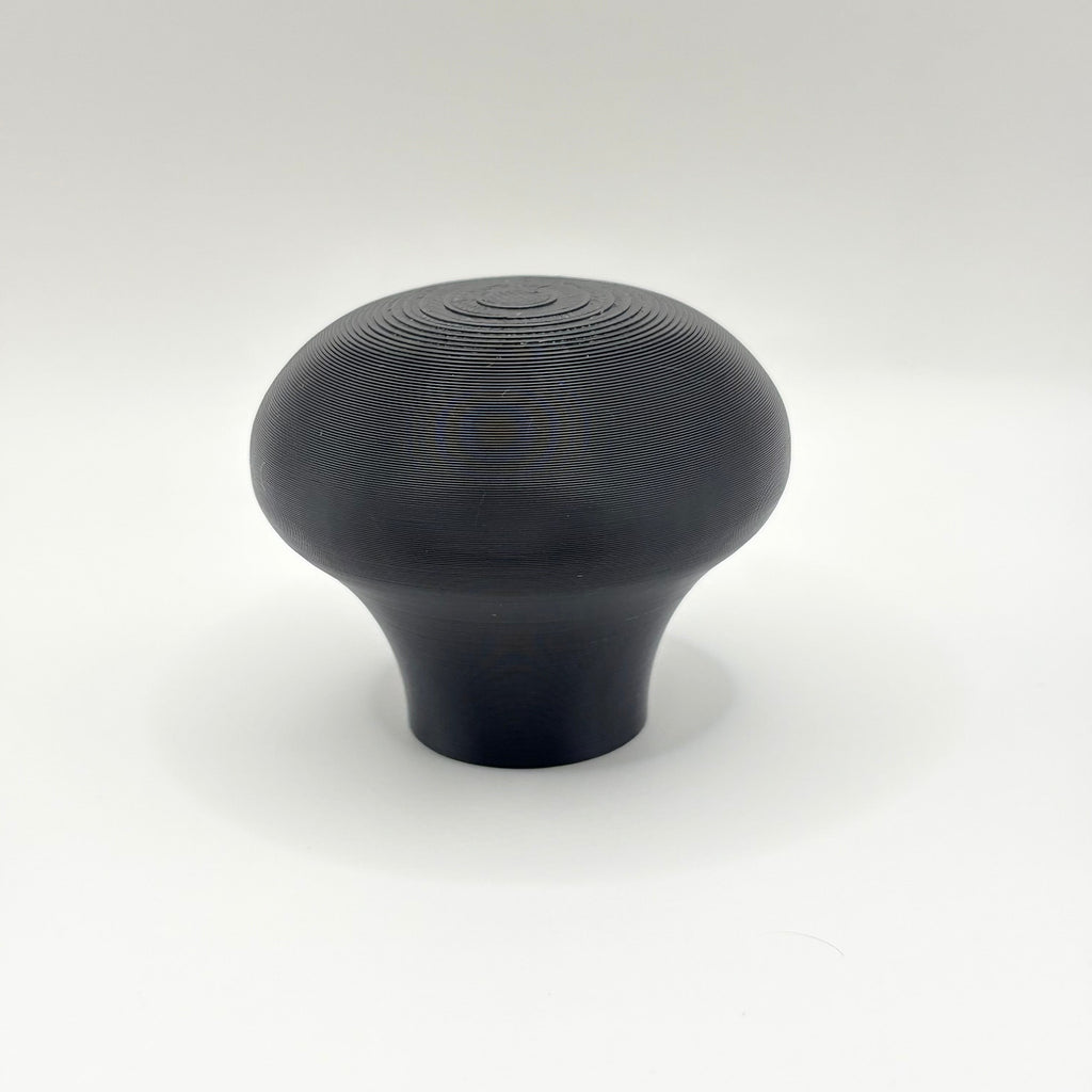 A single matte black knob or handle with a rounded, mushroom-like shape and subtle concentric ring texture on top, photographed against a clean white studio background.