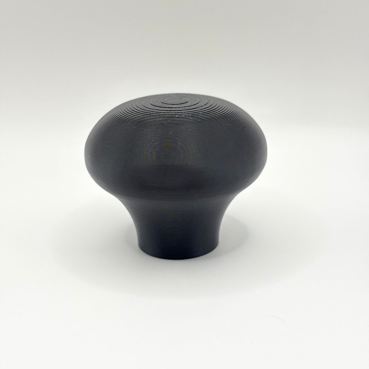 A single matte black knob or handle with a rounded, mushroom-like shape and subtle concentric ring texture on top, photographed against a clean white studio background.
