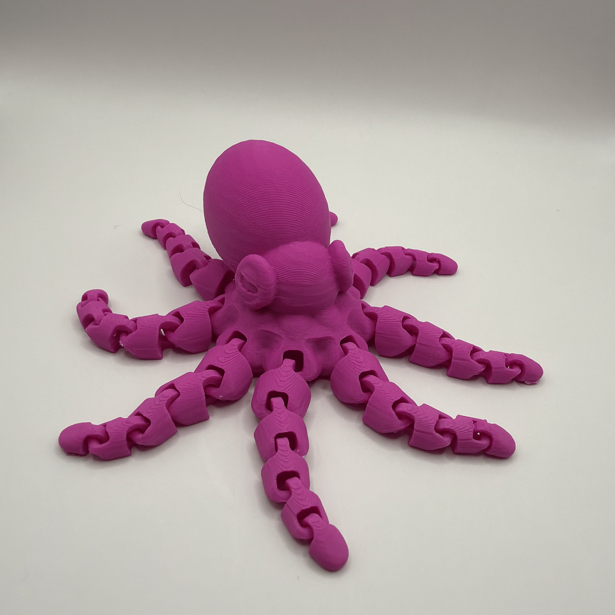 A bright magenta 3D-printed octopus toy with articulated, chain-link style tentacles and a smooth, rounded head, displayed on a neutral light gray background.