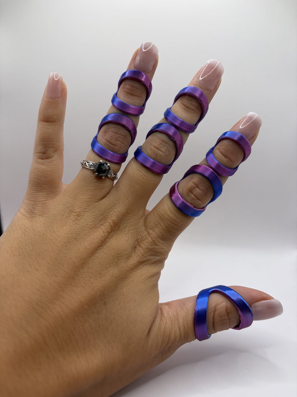 A hand wearing five purple and blue iridescent ring splints on the fingers and thumb, with a silver ring on the middle finger, against a plain white background.